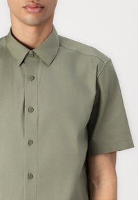 Chemise à manches courtes avec boutons en vert discret. Présente un col classique, une texture lisse et six boutons à l'avant pour la fermeture.
