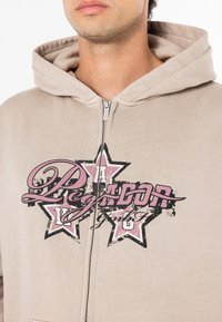 Beige zip-up hoodie with a large pink and black graphic design of stars and text on the front; cotton material, medium thickness.