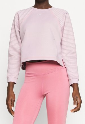 Personne portant un sweat matelassé rose clair à manches longues et un legging rose taille haute, debout sur un fond blanc.