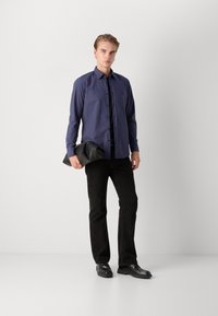Cămașă albastră cu butoni, blugi negri, ținând o geantă de mână din piele neagră. Material neted, croială obișnuită, mâneci lungi și tăietură dreaptă. Pantofi negri.