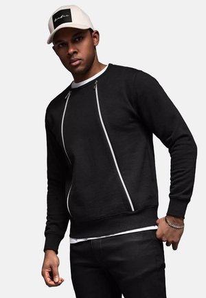 Svart sweatshirt med vita dragkedjedetaljer och rund halsringning, kombinerad med svarta byxor. En beige keps med svart logotyp fulländar looken.