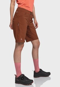 Braune Outdoor-Shorts mit tailliertem Schnitt, zwei Seitentaschen und Logo-Detail. Kombiniert mit schwarzen Schuhen und rosa Socken.