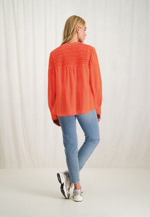 Oranje blouse met gerimpeld achterkantdetail, lange mouwen en geribbelde manchetten, gecombineerd met lichtblauwe jeans en sportieve sneakers.