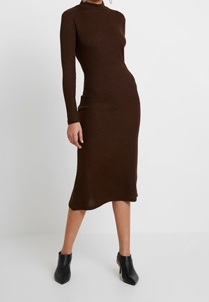 Woman wearing a fitted, long-sleeve, dark brown midi dress with a high neckline paired with black heeled ankle boots.