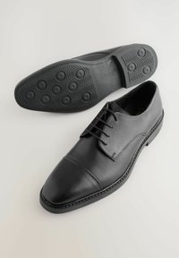 Zapato de vestir de cuero negro con punta redonda, diseño de cordones y suela de goma texturizada con tacos circulares para mayor agarre.