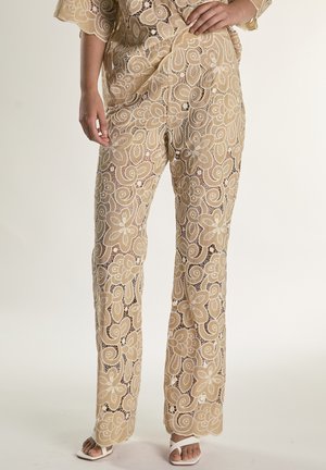 Beige kanten broek met bloemen- en spiraalpatronen, gedragen met bijpassende top en witte open-toe hakken, handen rustend op de heupen.