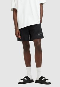 Black shorts made of lightweight fabric with a straight cut, featuring the logo "ALLSAINTS" in white. Worn with a loose white tee and black sandals.