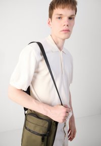 Sac bandoulière vert olive en tissu lisse, avec une poche avant zippée et une longue bandoulière noire. Le modèle porte un polo de couleur claire.