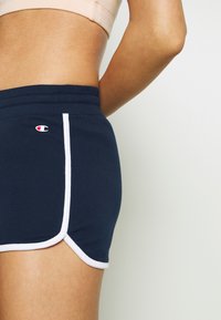 Kvinna som bär marinblå shorts med vitt kantband och en liten röd- och vit logotyp på sidan, visad från midjan till mitt på låret.