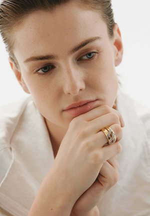 Anillo entrelazado de oro y plata con un diseño texturizado, llevado por una persona de piel clara que descansa suavemente su barbilla en la mano.