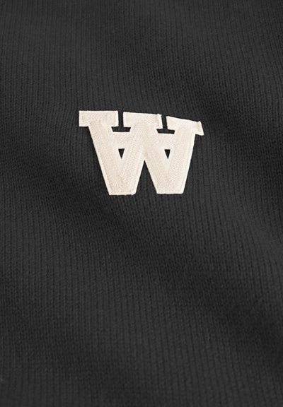 Tela de punto negra con una destacada letra "W" bordada en color beige con una tipografía estilizada. La textura es suave con un ligero patrón acanalado.
