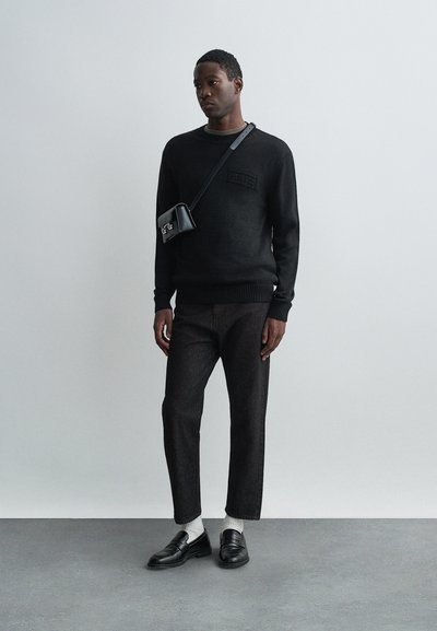 Vyras dėvi juodą megztinį, trumpas juodas kelnes, baltas kojines, juodus mokasinus ir mažą juodą per petį nešamą krepšį prieš pilką fono.