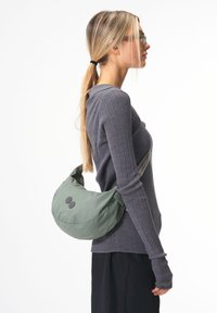 Grüne Nylon-Schultertasche mit einer weichen, abgerundeten Form. Verfügt über zwei schwarze Knöpfe und einen Riemen in einer kontrastierenden Farbe. Die Textur wirkt glatt.