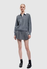 Szary sweter dziany w serek, z detalami w warkocze i ściągaczem na dole. Połączony z szarymi spódnicami plisowanymi oraz czarnymi loafersami.