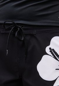 Shorts noirs avec une taille à cordon et un grand imprimé de fleur d'hibiscus blanche sur le côté droit.