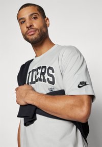 Pilkas trumpomis rankovėmis marškinėliai, kuriuose ryškiomis juodomis raidėmis užrašyta "RAIDERS", su juodu Nike logotipu ant rankovės ir tamsiai mėlyna megztiniu uždėtu ant rankos.