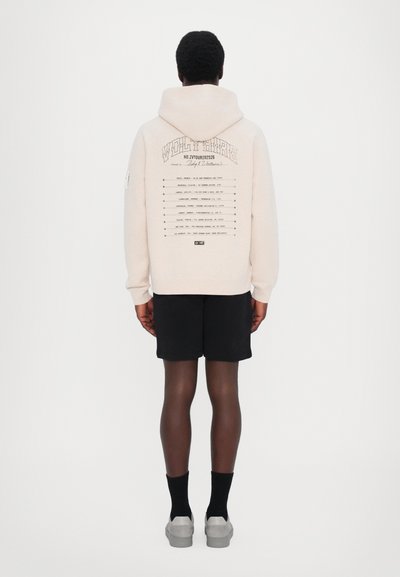 Pullover hoodie in light beige cotton with a large graphic on the back, featuring text and patterns. Paired with black shorts and shoes.