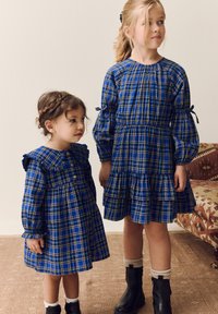 Due ragazze indossano vestiti blu a quadri, con maniche lunghe e dettagli arricciati. Il vestito della più giovane ha un colletto, e entrambi presentano gonne a balze.