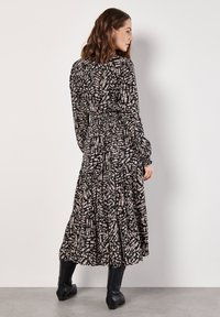 Schwarzes Kleid mit Muster in Maxilänge, langen Ärmeln, taillierter Taille und fließendem Rock. Die Textur wirkt weich und hat ein gestuftes Design. Kombiniert mit schwarzen Stiefeln.