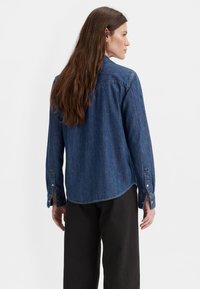 Chemise en denim bleu foncé à manches longues, avec des poignets à boutons et un ourlet arrondi. Équipée d'un col classique et d'un design uni sans motifs.