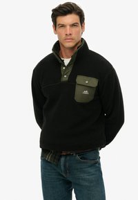 Sort fleece pullover med en kontrastfarvet olivengrøn krave og lomme. Har en kvart lynlås og et logo-patch på brystlommen.