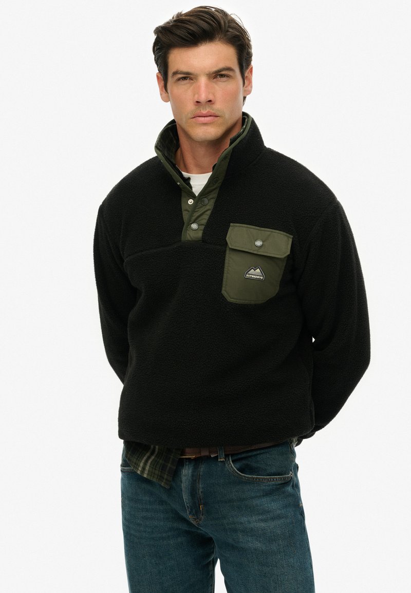 Sort fleece pullover med en kontrastfarvet olivengrøn krave og lomme. Har en kvart lynlås og et logo-patch på brystlommen.