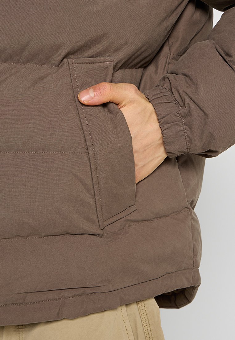 Giacca imbottita marrone con finitura testurizzata, dotata di una tasca laterale e polsini arricciati per una vestibilità aderente. Design semplice e funzionale.