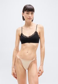 Γυναίκα που στέκεται φορώντας ένα μαύρο μπραλέτο με δαντέλα και μια μπεζ λεπτή εσώρουχο με δαντέλα, μπροστά σε απλό ανοιχτό φόντο.