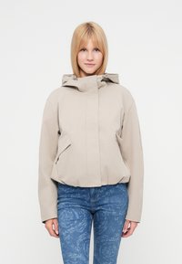 Μπεζ ζακέτα με κουκούλα σε χαλαρή γραμμή, με δύο εμπρόσθιες τσέπες και λείο υφή. Φορεμένη με μπλε τζιν με μοτίβο.