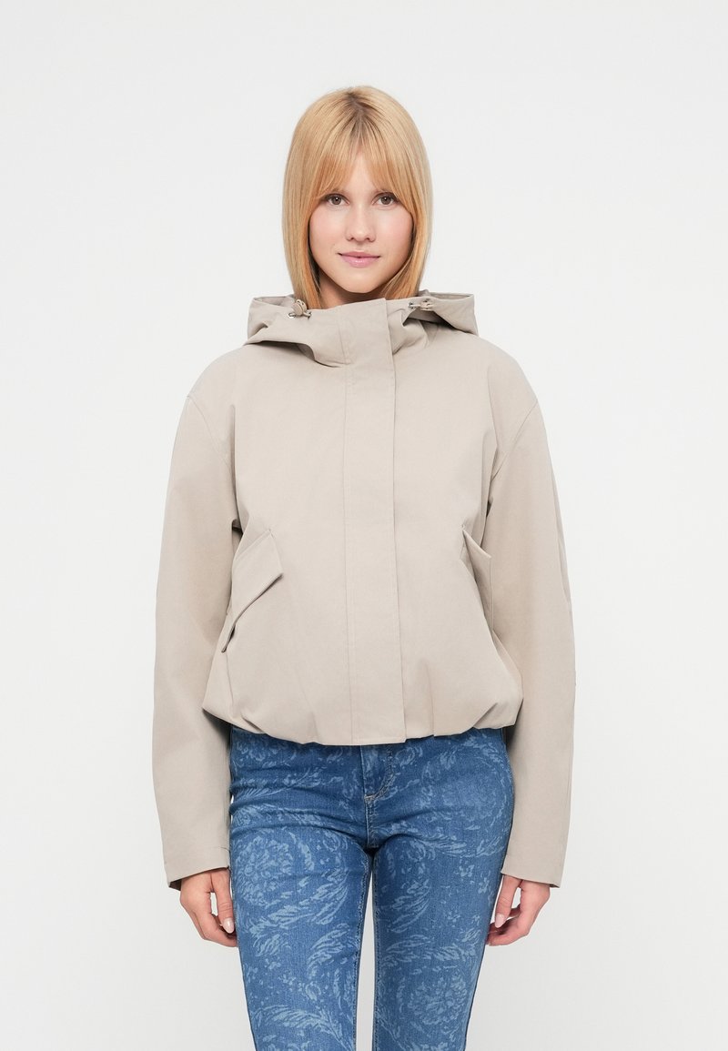 Μπεζ ζακέτα με κουκούλα σε χαλαρή γραμμή, με δύο εμπρόσθιες τσέπες και λείο υφή. Φορεμένη με μπλε τζιν με μοτίβο.