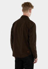 Brown corduroy shirt with a collar, long sleeves, and buttoned cuffs. Features a small red logo at the hem. Smooth texture.