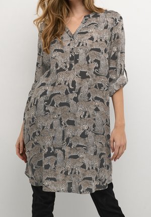 Femme portant une tunique à manches longues remontées, longueur genoux, avec un motif léopard noir et beige.