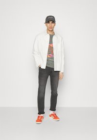 White denim jacket, gray graphic t-shirt with pink accents, dark jeans, and orange suede sneakers. Model wears a gray cap.