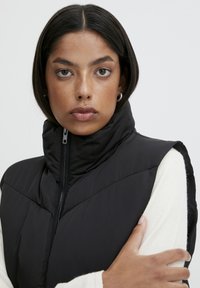 Gilet noir rembourré avec col haut, fermeture éclair et motif en chevrons. Porté par-dessus un haut à manches longues crème en tissu texturé.
