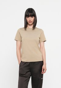 Beige short-sleeve t-shirt with a crew neckline and subtle logo stitching; worn with black satin trousers, standing against a white background.