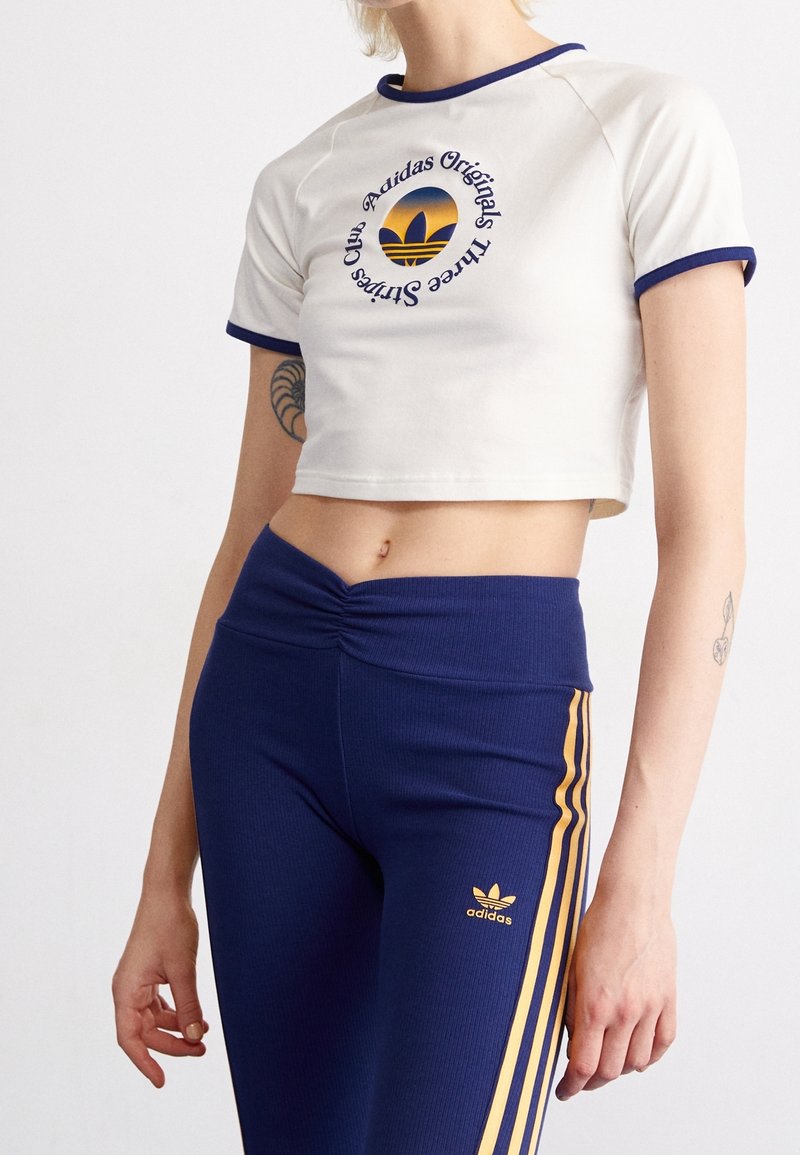 Bílý zkrácený tričko s modrým lemováním obsahující kulaté logo Adidas. Spárováno s přiléhavými námořnickými legínami se zlatými pruhy a logem.