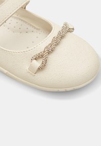 Weißer Kunstleder-Schuh mit strukturierten Oberflächen, versehen mit einer dekorativen verdrehten Akzentuierung und einem Riemen über dem Fußspann.