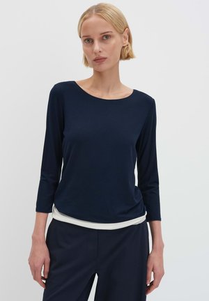 Marineblaues Langarmoberteil mit rundem Ausschnitt, weicher Textur und einem fallenden Design am Saum, kombiniert mit maßgeschneiderten marineblauen Hosen.