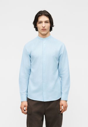 Jeune homme aux cheveux mi-longs foncés portant une chemise bleu clair à col mandarin boutonnée et un pantalon marron foncé.