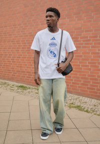 Hvid t-shirt med blåt Real Madrid-logo og tre blå striber fra Adidas; lyseblå baggy jeans; sort crossbody taske; marineblå sneakers.