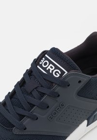 Marinblå idrottssko med nätövre och gummidetaljer. Har en framträdande "BORG"-logotyp, runda snören och perforerade sidodetaljer.