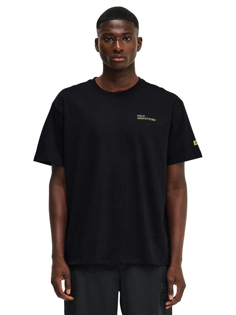 Mężczyzna w czarnej, oversize'owej koszulce z małym, żółtym napisem "HALO 196819721981" na piersi oraz żółtą łatą na lewym rękawie.