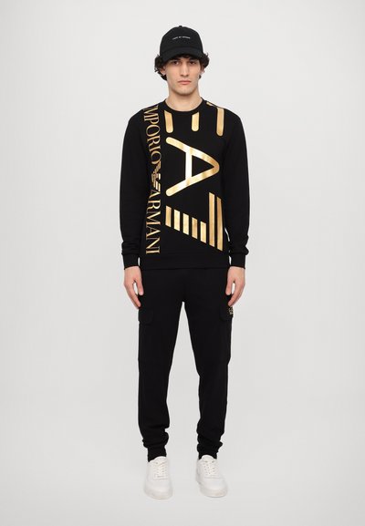 Sweatshirt noir avec texte graphique doré et accents rayés, associé à un pantalon cargo noir. Le modèle porte des baskets blanches et une casquette noire.