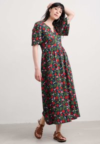 Robe maxi florale en vert foncé avec des fleurs rouges, roses et blanches. Décolletée en V, manches courtes bouffantes, taille froncée, longueur jusqu'aux chevilles avec une coupe fluide. Sandales brunes à lanières.