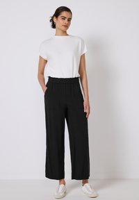 Haut blanc à manches courtes associé à un pantalon large noir avec taille élastique et poches. Le modèle porte des baskets blanches.