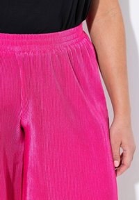 Pantaloni plissettati rosa con una finitura testurizzata, dotati di una ampia vita elastica e un design fluente. Primo piano dei dettagli del tessuto.