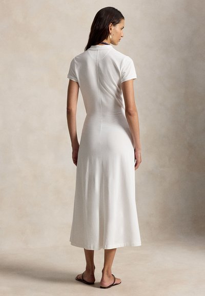 Hosszú, sötét hajú nő, fehér, rövid ujjú midi ruhát és fekete szandált viselve, háttal áll egy semleges hátterű térben.