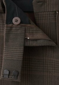 Brązowa i ciemna tkanina w kratę z guzikiem, zamkiem błyskawicznym i dwoma czarnymi plastikowymi haczykami na krawędzi odzieży, ukazująca szczegółową fakturę i ścieg.