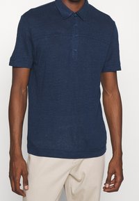 Muž oblečený v polo tričku s krátkým rukávem námořnické modré barvy s knoflíky a světle béžových kalhotách, zobrazený od krku po boky.