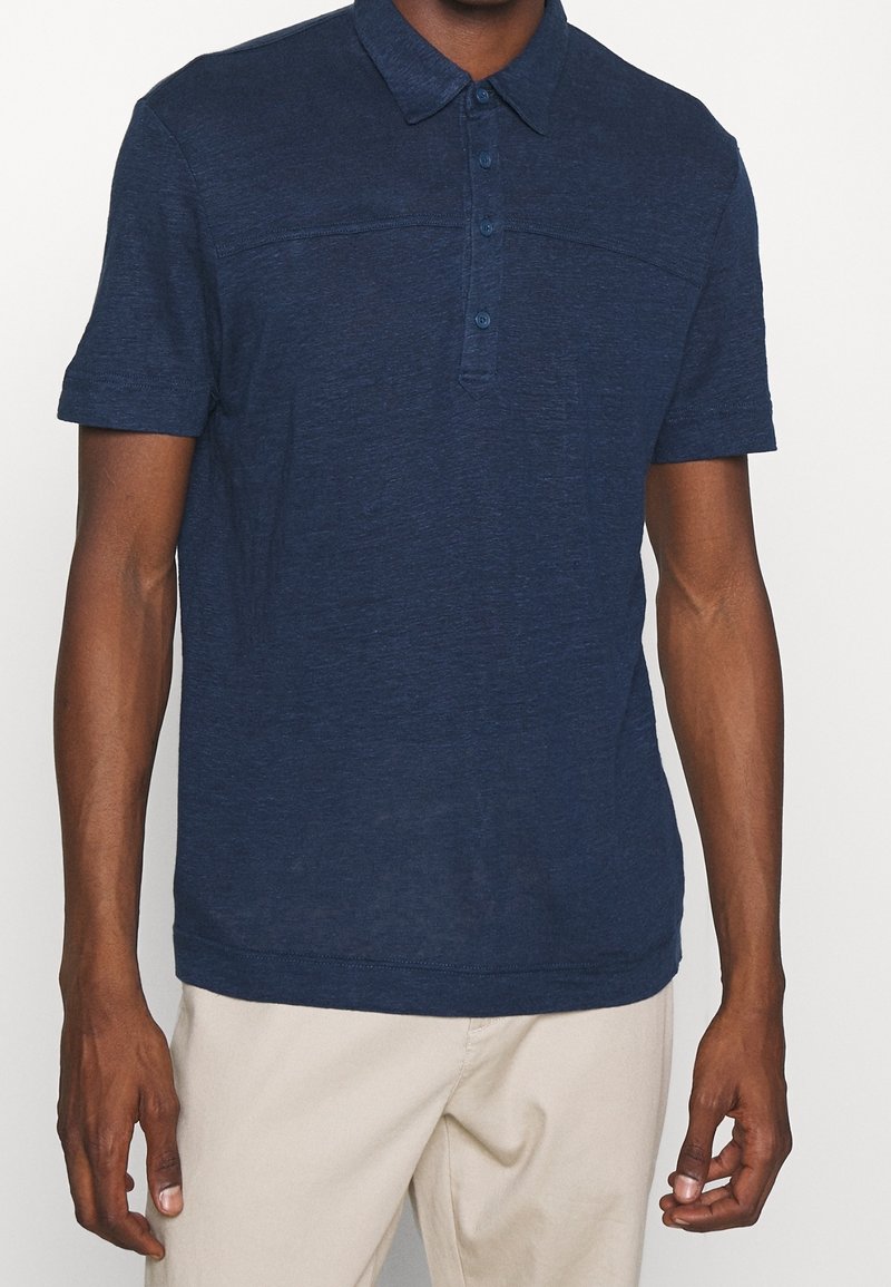 Muž oblečený v polo tričku s krátkým rukávem námořnické modré barvy s knoflíky a světle béžových kalhotách, zobrazený od krku po boky.